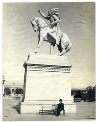 Verticaal, sepiafoto van een standbeeld van een Amerikaanse Indiaan zittend op een paard op een groot voetstuk. Er zit een man in een donker pak en hoed op een bankje aan de voet van het voetstuk
